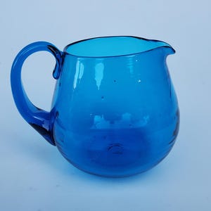 May include: A vibrant blue glass pitcher with a rounded body and a curved handle. The pitcher has a spout for easy pouring. The glass is transparent, allowing light to pass through. This decorative pitcher is suitable for serving beverages.