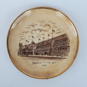 May include: A brown and white ceramic plate with a gold rim. The plate features a black and white illustration of a building with the text "Clay Products Exposition The Coliseum Chicago" and "March 7th to 12th 1912".