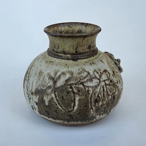 May include: A handcrafted ceramic vase with a round body and a flared neck. The vase features a textured surface with a floral pattern in shades of brown and green. It has a small handle on the side and is approximately 7 inches tall.