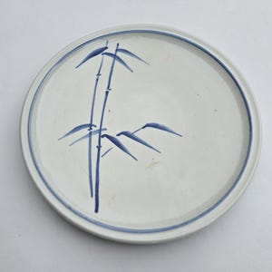 May include: White ceramic plate with a blue bamboo design. The plate has a blue rim and a hand-painted bamboo stalk and leaves in the center. The plate is round and appears to be about 10 inches in diameter.