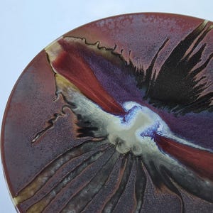 May include: A circular ceramic plate featuring a glossy glaze with a blend of deep burgundy, black, and cream colors. The surface displays a radiating pattern with dark streaks extending from a central cream-colored area toward the outer edge.