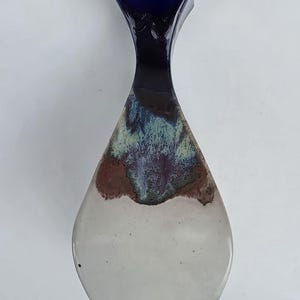 May include: A handcrafted ceramic vase with a unique shape and color gradient. The top is a deep blue, transitioning through shades of brown, green, and purple, to a white base. The vase is approximately 10 inches tall.