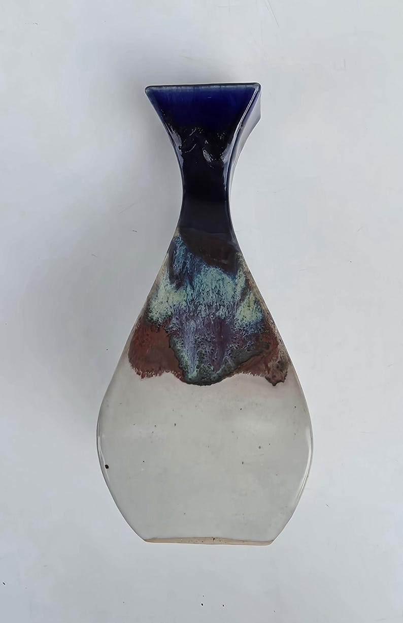 May include: A handcrafted ceramic vase with a unique shape and color gradient. The top is a deep blue, transitioning through shades of brown, green, and purple, to a white base. The vase is approximately 10 inches tall.
