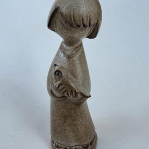 May include: A ceramic figurine of a stylized figure with a rounded head and a long dress. The figure is holding a bird. The figurine is a speckled brown color and has a textured surface. The base of the figurine has a decorative border.