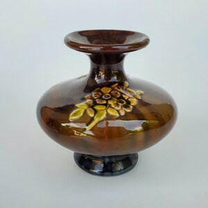 May include: A brown ceramic vase with a wide base and a narrow neck. The vase has a raised floral design in shades of green, yellow, and brown.