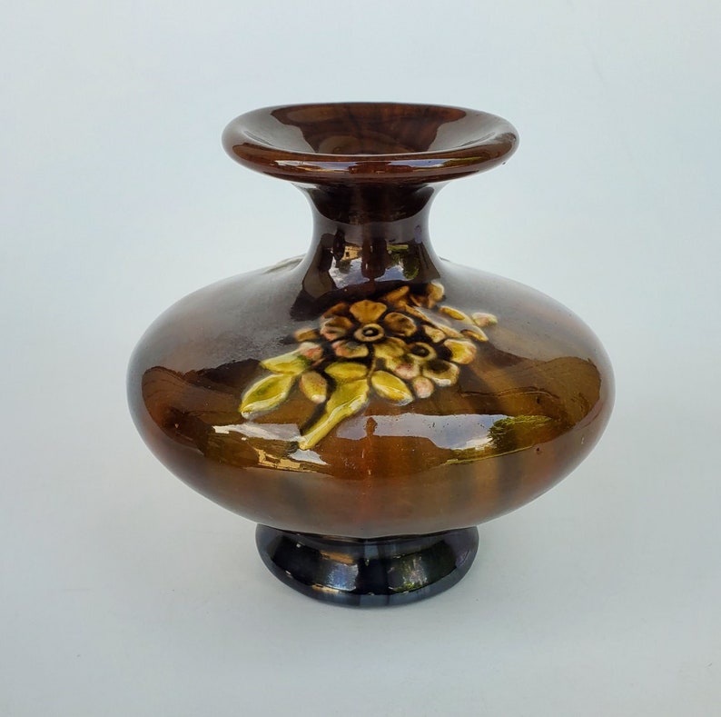 May include: A brown ceramic vase with a wide base and a narrow neck. The vase has a raised floral design in shades of green, yellow, and brown.