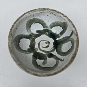 May include: A round ceramic bowl with a speckled off-white glaze and a dark green hand-painted floral design at the centre. The bowl features a thick, textured rim with a mottled brown and grey finish, showing visible brushstroke details.