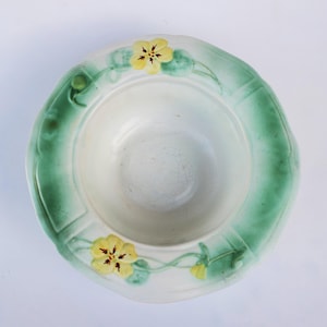 May include: A white ceramic bowl with a green and yellow floral design. The bowl has a scalloped edge and is decorated with stylized flowers and leaves.