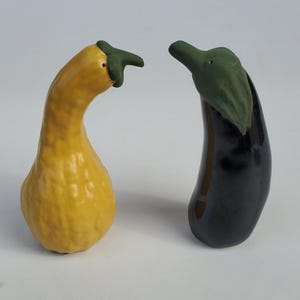 May include: A set of ceramic salt and pepper shakers shaped like a yellow squash and a black eggplant. The squash has a green stem and the eggplant has a green leafy top.