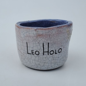 May include: A white ceramic pot with a crackled glaze and the inscription "Leo Holo" in black ink.