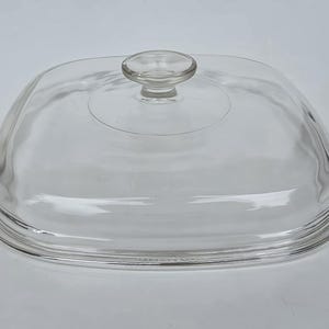May include: A clear, square-shaped glass lid with rounded corners and a centered, circular knob handle. The lid features a slightly domed top surface and a stepped rim design, providing a transparent cover for a square cooking vessel or dish.