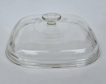 May include: A clear, square-shaped glass lid with rounded corners and a centered, circular knob handle. The lid features a slightly domed top surface and a stepped rim design, providing a transparent cover for a square cooking vessel or dish.