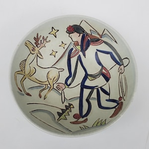 May include: A ceramic bowl with a hand-painted design of a person in a blue and white outfit with a red hat, holding a rope and walking with a deer in a snowy landscape. The bowl has a white background with blue, red, yellow, and green accents.