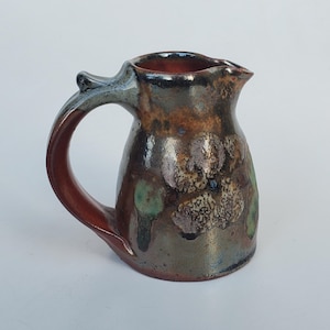 May include: A handmade ceramic pitcher with a unique, textured glaze. The pitcher features a dark brown and green mottled pattern with a reddish-brown handle. The interior is a deep red color. The pitcher has a spout for easy pouring.