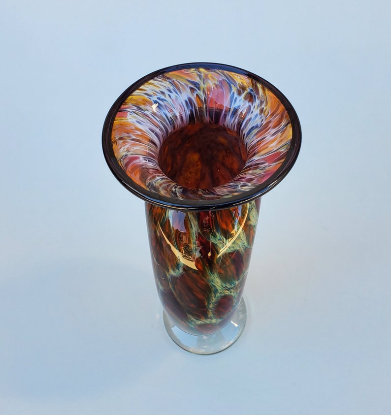 May include: A tall, brown glass vase with a wide, flared opening. The vase has a swirling pattern of white, orange, and red colours. The opening of the vase has a black rim.