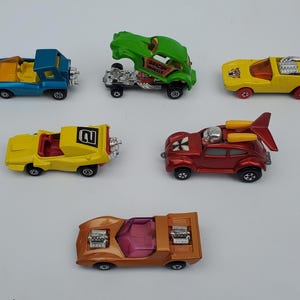 May include: A collection of six toy cars, including a red Volkswagen Beetle with a plane on top, a yellow car with a number 2 on the side, a blue car with a yellow stripe, a green car with a dragon design, a yellow car with a red stripe, and an orange car with a pink interior.