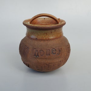 May include: A brown ceramic honey pot with a lid and a handle. The pot has the word "honey" inscribed on the side.