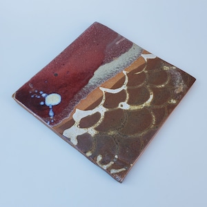 May include: A square ceramic tile with a brown, red, and white abstract design. The tile has a textured surface and a unique pattern.