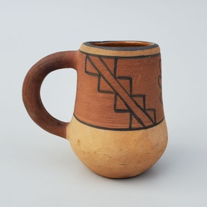 May include: Handmade ceramic mug with a rounded base and a dark brown handle. The upper portion of the mug is a reddish-brown color, decorated with black geometric designs. The lower portion is a lighter tan color. The mug is suitable for hot or cold beverages.