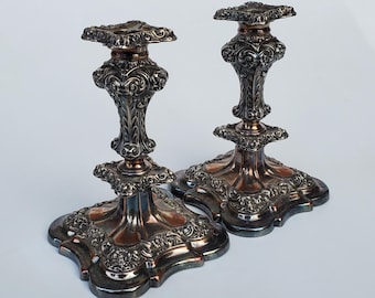 May include: Two antique silver candlesticks with ornate, floral designs. The candlesticks have a square base and top, with a detailed, curved stem. The silver has a tarnished appearance, adding to their vintage charm.