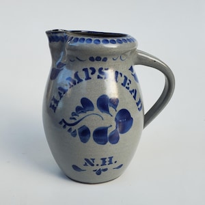 May include: A gray ceramic pitcher with a handle and spout. The pitcher is decorated with blue floral designs and the word "Hampstead" in blue lettering. The letters "N.H." are also visible. The pitcher has a classic, rustic appearance.