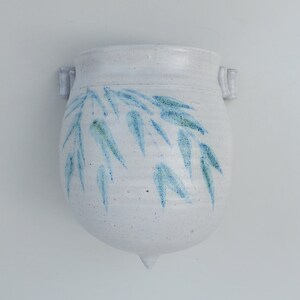 May include: White ceramic wall planter with a blue and green leaf design. The planter has two small handles on the sides and a pointed bottom.