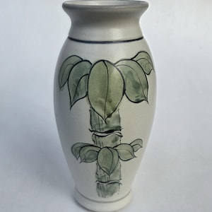 May include: A cream-colored ceramic vase with a flared rim and a tapered base. The exterior features a hand-painted green bamboo stalk design with detailed leaves and dark outlines. The surface has a smooth, matte finish with a thin dark line.