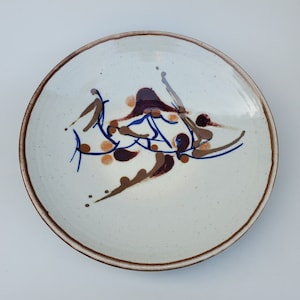 May include: A white ceramic plate with a brown and blue abstract design. The design features swirling lines and dots.