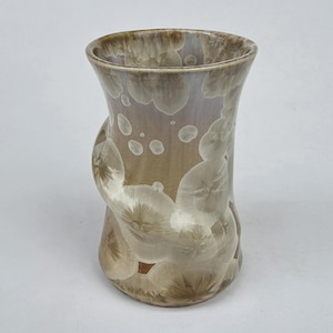 May include: A ceramic vase with a flared rim and a smooth, glossy finish. The surface features a crystalline glaze pattern with overlapping, petal-like shapes in shades of beige, taupe, and soft white, creating a layered, organic visual effect.