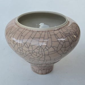 May include: A pale pink ceramic bowl with a crackle glaze pattern. The bowl has a wide, open top and a rounded body that tapers to a small, pedestal base. The crackle pattern is a network of fine, dark lines across the surface.