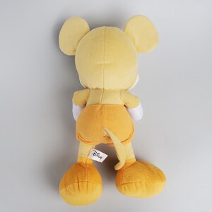 Puede incluir: Un juguete de peluche Mickey Mouse amarillo con una etiqueta blanca del logotipo de Disney. El juguete est&aacute; de pie sobre un fondo blanco.