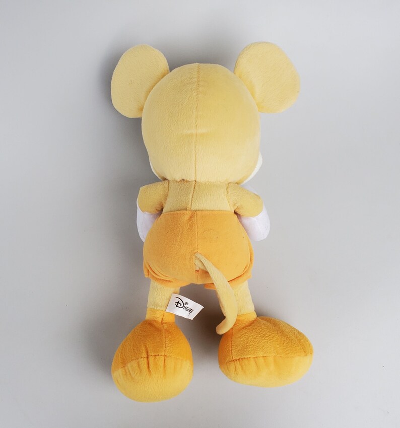 Puede incluir: Un juguete de peluche Mickey Mouse amarillo con una etiqueta blanca del logotipo de Disney. El juguete est&aacute; de pie sobre un fondo blanco.