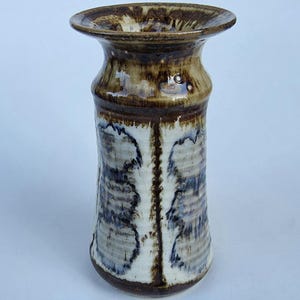 May include: A handcrafted ceramic vase with a wide, flared rim and a cylindrical body. The vase features a color palette of brown, white, and blue, with a textured, glazed finish. The design includes vertical patterns and a dark brown base.