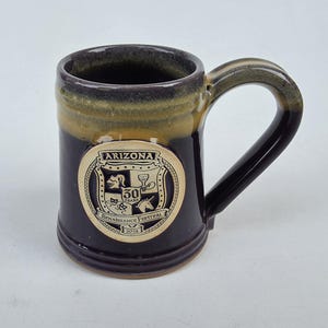 May include: A ceramic mug with a dark brown exterior and a tan rim. The mug features a circular emblem with the text "ARIZONA Renaissance Festival 50 Years 2018". The handle is dark brown.