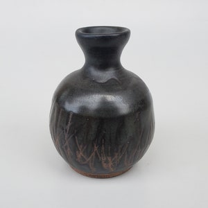 May include: A small, dark gray ceramic vase with a textured, brown and black design. The vase has a round body and a narrow neck.