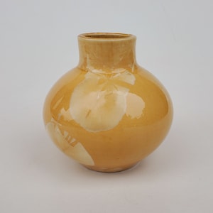 May include: A small, round, yellow ceramic vase with a white glaze design. The vase has a wide mouth and a narrow neck.