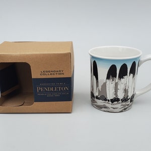 May include: A white ceramic mug with a black and white feather design. The mug has a blue sky background and the text "WARRANTED TO BE A LEGENDARY COLLECTION PENDLETON PENDLETON WOOLEN MILLS SINCE 1882" on the box.