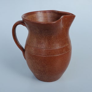 May include: A terracotta pitcher with a wide mouth and a handle. The pitcher has a rustic, hand-thrown look and is glazed in a warm brown color.