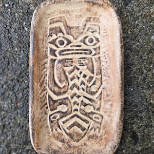 May include: A rectangular, ceramic trinket dish with a carved totem pole design. The dish is a light brown color with darker brown detailing in the carved design. The dish is approximately 5 inches long and 3 inches wide.
