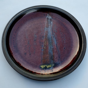 May include: A round ceramic plate with a dark brown rim and a red glaze. The glaze has a metallic sheen and a vertical streak of gold and white.