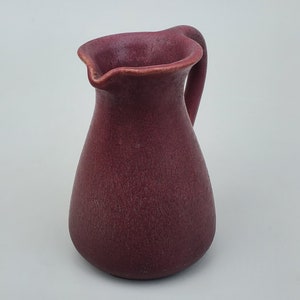 May include: A small, burgundy ceramic jug with a rounded body and a slightly flared lip. The jug has a small handle on the side.
