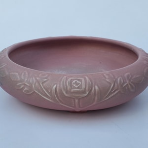 May include: A dusty rose-colored ceramic bowl with a low profile and a wide rim. The rim features a repeating floral pattern in a lighter shade, with a central rose motif. The bowl is likely for decorative or functional use.