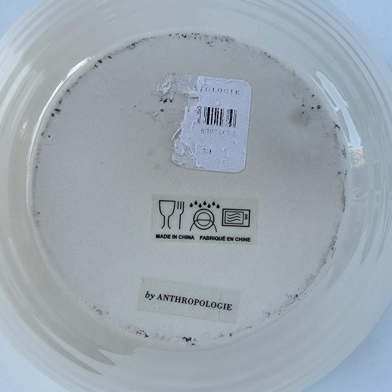 Peut inclure: Assiette en c&eacute;ramique blanc cass&eacute; avec un bord stri&eacute;. Le dessous de l'assiette comporte une &eacute;tiquette avec un code-barres et le texte "by ANTHROPOLOGIE". L'assiette comporte &eacute;galement des symboles indiquant qu'elle est lavable au lave-vaisselle et au micro-ondes.