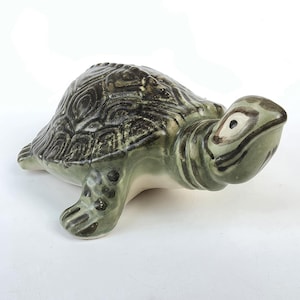 May include: A ceramic turtle figurine featuring a glossy finish in shades of sage green and dark olive. The shell displays a detailed, raised geometric pattern, while the head and limbs are marked with dark green spots and lines on a lighter base.