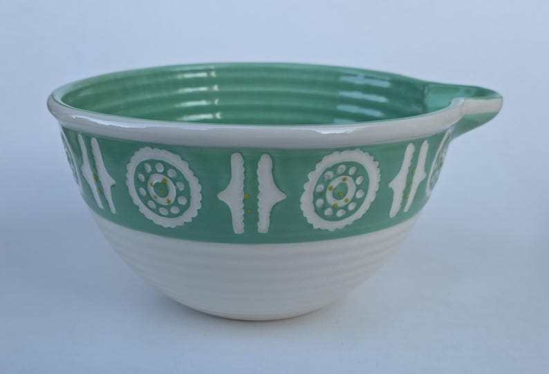 Peut inclure: Un saladier en c&eacute;ramique blanc et turquoise. Le bol a une poign&eacute;e et une bande d&eacute;corative avec des motifs circulaires et lin&eacute;aires blancs et jaunes. La moiti&eacute; inf&eacute;rieure du bol est blanche et le haut est turquoise.