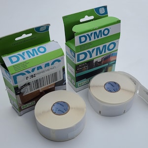Peut inclure: Deux rouleaux de ruban d'étiquettes DYMO et deux boîtes vertes et blanches pour étiqueteuse DYMO. Les boîtes sont ouvertes et les rouleaux d'étiquettes sont blanc cassé. Les boîtes portent le logo DYMO et des informations sur le produit.
