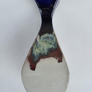 May include: A ceramic vase with a unique shape and a glossy finish. The top is a deep blue, transitioning to a dark brown and green midsection, and a white base. The vase is a decorative item.