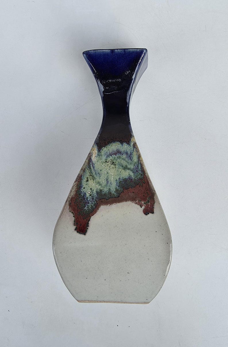 May include: A ceramic vase with a unique shape and a glossy finish. The top is a deep blue, transitioning to a dark brown and green midsection, and a white base. The vase is a decorative item.