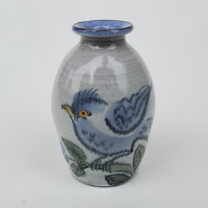 May include: A small, gray ceramic vase with a blue rim and a hand-painted design of a blue bird with yellow eyes and green leaves.
