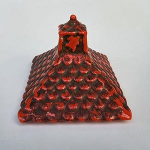 May include: A square ceramic lid featuring a raised, textured roof tile pattern in a vibrant orange and black finish. The lid is topped with a small, square chimney-like handle, showcasing a glossy glaze with dark distressed detailing.